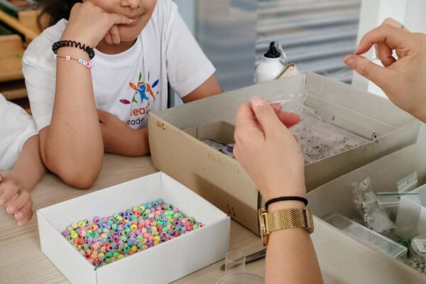 Mädchen und Erwachsene basteln bunte Perlenarmbänder. T-Shirt mit "Learner Academy" Logo.