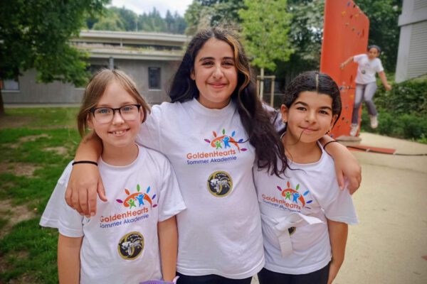 Drei lächelnde Mädchen in GoldenHearts Sommer Akademie T-Shirts, eines klettert im Hintergrund.