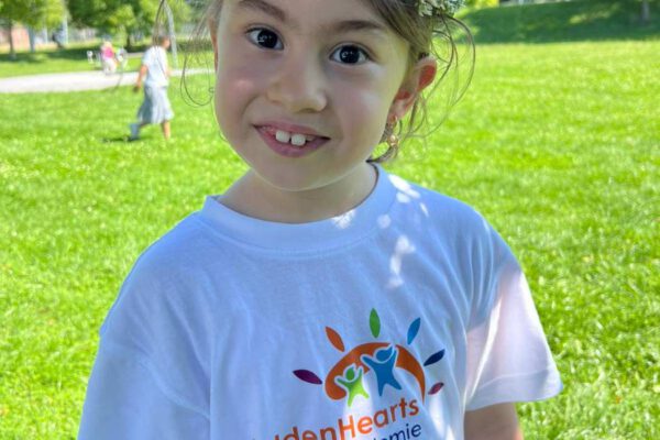Lachendes Mädchen mit Blumenkranz, GoldenHearts Sommer Akademie T-Shirt & Tavido Zentrum Logo im Park.